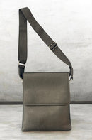 A black pebbled leather crossbody bag with a wide adjustable shoulder strap, standing upright against a textured gray concrete wall. By Fresh N Fitted.