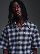 A man with long black dreadlocks wearing a short-sleeve plaid button-up shirt with 'GUAPI' printed in white on the chest, photographed against a dark gray studio background. By fresh N Fitted.