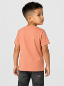A young boy wearing a solid peach-colored short-sleeve t-shirt and dark gray jeans, photographed from the back against a light gray studio background.