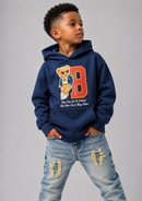 A young boy wearing a navy blue hoodie with a cartoon teddy bear graphic and a large orange number 8 on the front, paired with distressed light blue jeans, standing against a solid gray studio background.