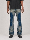 A person wearing light-wash blue distressed jeans with large dark blue patch panels on the thighs, shown from the waist down against a light gray studio background.
