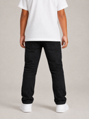 A child shown from the back wearing black denim jeans, a white t-shirt, and white sneakers against a neutral studio background.