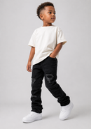 A young boy modeling a casual outfit consisting of an oversized off-white t-shirt, black distressed jeans, and white sneakers, photographed against a neutral light gray studio background.