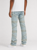 A person shown from the back wearing light-wash blue denim jeans with diagonal patchwork paneling and white sneakers against a plain light gray studio background.