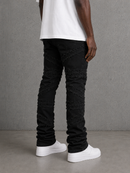 A person shown from the waist down wearing black distressed jeans with textured, shredded detailing running vertically and diagonally down the legs, styled with a white t-shirt and white platform sneakers against a gray concrete wall.