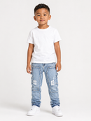 A young boy modeling a white crewneck t-shirt, light wash blue distressed jeans, and white sneakers against a plain white studio background.