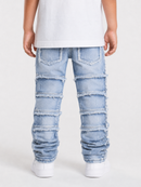A child viewed from the back wearing light-wash blue distressed denim jeans with layered ripped panel details and frayed edges, paired with a white t-shirt and white sneakers against a light gray studio background.