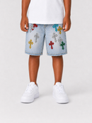 A child wearing light-wash denim shorts with colorful embroidered crosses in red, yellow, green, and silver, paired with a white t-shirt and white sneakers, shown from the waist down on a light gray studio background.