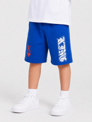 A child wearing bright royal blue shorts with a large white distressed graphic on the right leg and a small red heart-and-letter graphic on the left leg, paired with a white shirt, white socks, and white sneakers on a light gray studio background.
