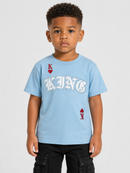 A young boy wearing a light blue graphic t-shirt with a white gothic 'KING' text and red heart suit graphics, paired with black cargo pants, against a light gray background.