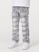Light gray heavily distressed denim jeans with ripped paneling and frayed edges, shown on a model from the waist down wearing white sneakers against a light gray studio background.