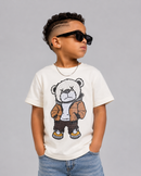 A child wearing a cream-colored t-shirt with a large teddy bear graphic, styled with blue jeans, black sunglasses, and a silver chain necklace against a solid gray background.