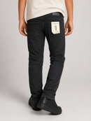Rear view of a person wearing slim-fit black jeans with a visible back pocket patch and black shoes, standing against a light gray studio background.