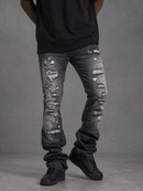 Distressed black and gray washed denim jeans with ripped details, worn by a model in a black t-shirt and black sneakers against a gray studio background.