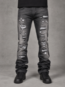 Distressed black denim jeans with heavy rips and frayed detailing on the legs, shown on a model from the waist down wearing black sneakers against a gray studio background.