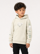 A young boy wearing a light beige hoodie with green and white graphic prints on the chest and sleeves, paired with black pants, photographed against a plain white studio background.