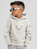 A young boy wearing a light beige Givenchy hoodie with graphic prints on the front and sleeves, styled with white sunglasses and light wash jeans against a solid light gray background.