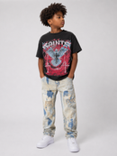 A child model wearing a black graphic t-shirt with a red and blue Saints band print, paired with distressed light-wash jeans and white sneakers, standing against a light gray studio background. By Fresh N Fitted