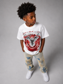 A young boy with curly black hair stands against a gray studio background wearing a white t-shirt with a red, black, and gray eagle graphic and the word 'SPITfires', paired with distressed beige and blue paint-splattered jeans and white sneakers. By Fresh N Fitted