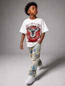 A child model wearing a white graphic t-shirt with a red and gray eagle design, distressed light-wash jeans with blue paint splatters, and white sneakers, standing against a gray studio background. By Fresh N Fitted