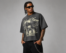 A man modeling an oversized dark gray graphic t-shirt with a cream-colored print that reads 'Silent Verse' and 'FAITH FOUND IN SILENCE'. He is also wearing black sunglasses, a gold chain necklace, and black pants against a neutral gray studio background. By Fresh N Fitted