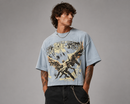 A man wearing a light blue graphic t-shirt with a 'Silent Warfare' angel print, styled with black jeans and a silver chain necklace against a gray studio background. By Fresh N Fitted