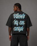A man shown from the back wearing a black oversized t-shirt with a large graphic print on the back in light blue and white gothic lettering that reads 'Raised by an angel'. By Fresh N Fitted