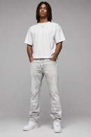 A man modeling a white crewneck t-shirt, light-wash gray jeans, and white sneakers against a solid gray studio background. By Fresh N Fitted