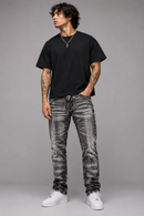 A man modeling a black crewneck t-shirt and distressed black-and-white acid-wash jeans with white sneakers, standing against a solid gray studio background. By Fresh N Fitted