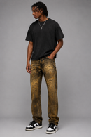 A man modeling a pair of brown acid-wash jeans with a black t-shirt and black-and-white sneakers, standing against a solid gray studio background. By Fresh N Fitted