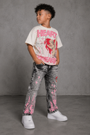 A child model wearing a natural beige graphic t-shirt with pink text and a red heart illustration, paired with gray acid-wash jeans featuring pink flame and star patterns, and white sneakers, standing against a textured gray wall. By Fresh N Fitted.