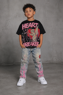A young boy stands against a textured gray studio wall wearing a black graphic T-shirt with pink 'HEART BREAKER' text and a red angel illustration, light-wash distressed jeans with pink splatter details, and white sneakers. by Fresh N Fitted