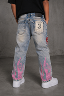 A person viewed from the back wearing light-tint jeans with bright pink flame graphics near the hems, a black shirt, and white sneakers against a gray studio backdrop. By Fresh N Fitted.