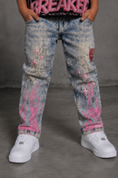 A child wearing light tint acid-wash jeans with pink graffiti-style paint splatters, black script embroidery, and a red heart patch, paired with white sneakers. By Fresh N Fitted.