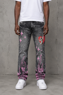 A person wearing washed black acid-wash jeans with pink flame and star graphics, a red heart patch, and a white t-shirt and white sneakers, standing against a gray concrete wall. By Fresh N Fitted.
