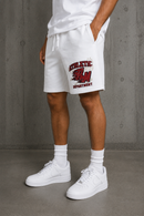 White athletic sweat shorts with a red and black 'Athletic Department' graphic on the left leg, worn by a model with white crew socks and white sneakers against a gray concrete wall. By Fresh N Fitted 