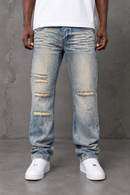 A person wearing light tint distressed denim jeans with ripped details on the thighs and knees, paired with a white t-shirt and white sneakers, standing against a gray textured wall. By Fresh N Fitted.