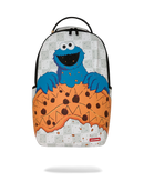 A backpack featuring a graphic print of the Cookie Monster character emerging from a giant chocolate chip cookie, with a gray newspaper pattern background and a black top handle.
