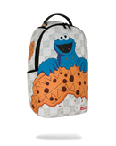 A Sprayground Cookie Monster backpack featuring a graphic print of Cookie Monster holding a giant chocolate chip cookie, set against a light gray patterned background.