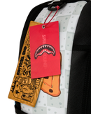 A close-up of a backpack showing its white and gray patterned fabric, black straps, and two hanging tags: a red tag with a shark-mouth graphic and vertical SPRYGROUND text, and a tan tag with cartoon graphics and the text SPRYGROUND.