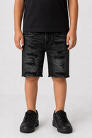 A child wearing distressed black denim shorts with a raw hem, paired with a black t-shirt and black sneakers, shown from the waist down against a light gray studio background.