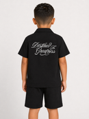 A child model shown from the back wearing a black short-sleeve collared shirt and matching black shorts. The back of the shirt features white cursive text reading 'Destined For Greatness'.