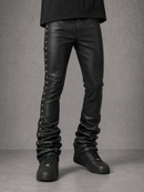 Black leather skinny jeans with decorative silver studs running down the outer sides, shown on a model from the waist down wearing black sneakers against a gray studio background.