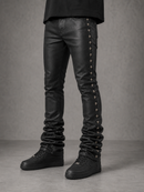 Black leather skinny jeans with decorative silver-tone cross studs down the outer side of each leg, shown on a model from the waist down against a textured gray studio background.