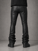 Rear view of a person wearing black leather pants with decorative side studs and gathered detailing at the lower legs, paired with black shoes against a gray studio background.