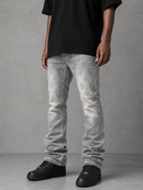 A person wearing light gray washed denim jeans, a black t-shirt, and black sneakers, standing against a textured gray studio background.