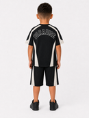 A child model viewed from the back wearing a black athletic jersey and matching shorts with white side panels and the word 'PARADISE' in white outlined lettering across the upper back, paired with black sneakers on a light gray studio background.