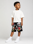 A young boy modeling a white crewneck t-shirt and black shorts featuring colorful skull and rose graphics, paired with white socks and sneakers, against a light gray studio background.