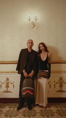 A man and a woman pose together in an ornate room, each carrying a shark-mouth patterned shoulder bag. The man wears a black blazer and shirt, and the woman wears a black top and long cream skirt.