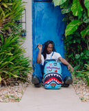 A man sitting on a concrete path in front of a blue door, wearing a white t-shirt, blue jeans, and holding a black object. In front of him is a light blue shark-mouth backpack decorated with colorful graphics and stickers.